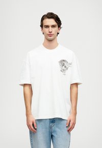 Λευκό βαμβακερό μπλ�ουζάκι με γραφιστικό σχέδιο στη αριστερή πλευρά του στήθους, χαλαρή εφαρμογή και κοντά μανίκια, σε συνδυασμό με ανοιχτό μπλε τζιν.