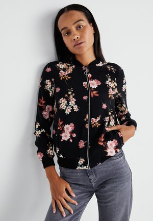 Veste bomber florale noire, ornée de fleurs roses et blanches, avec fermeture éclair et poignets et ourlet côtelés ; associée à un jean gris.