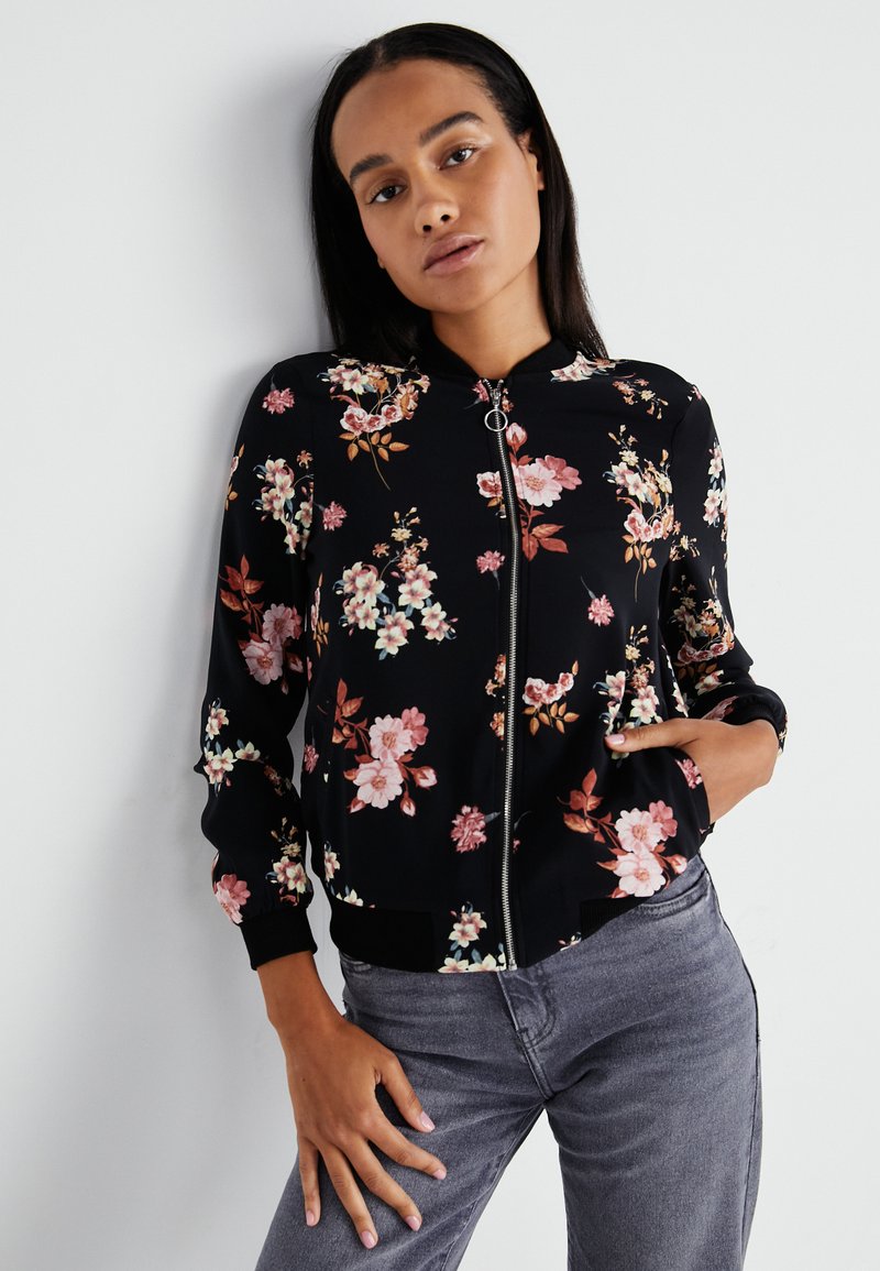 Veste bomber florale noire, ornée de fleurs roses et blanches, avec fermeture éclair et poignets et ourlet côtelés ; associée à un jean gris.