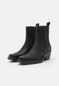 Botas de tobillo de piel negra con superficie texturizada, punta afilada y paneles laterales elásticos. Presentan un grueso tacón cuadrado y un diseño minimalista.