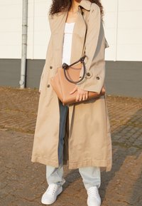Trench-coat beige avec un design à boutons, associé à un jean bleu clair et des baskets blanches, tenant un sac à main en cuir marron clair avec une poignée noire.