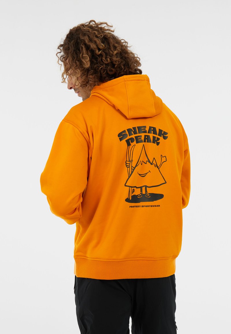 Mann mit lockigem Haar, der einen orangenen Hoodie trägt, auf dem ein Bergcharakter zu sehen ist, der Skier hält, und der Text "SNEAK PEAK" auf dem Rücken.