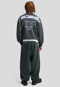 Temno siva oversize pula z belim grafičnim besedilom na hrbtni strani, v kombinaciji s širokimi temnimi hlačami. Teksturno blago in kontrastne šive.