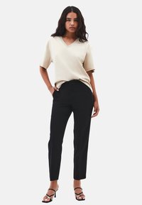 Mujer vistiendo una blusa beige de cuello en V y mangas cortas, pantalones negros ajustados y sandalias de tacón negras, de pie con una mano en el bolsillo.