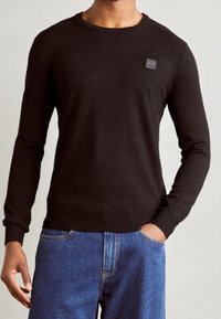 Czarny sweter z okrągłym dekoltem, długimi rękawami, ściągaczami na mankietach i u dołu, z kwadratową naszywką z logo na piersi, wykonany z gładkiego materiału.