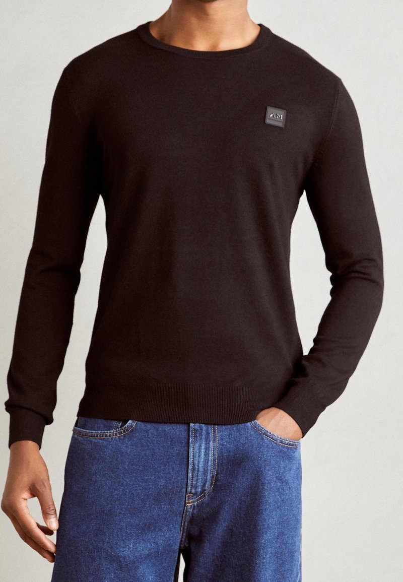 Czarny sweter z okrągłym dekoltem, długimi rękawami, ściągaczami na mankietach i u dołu, z kwadratową naszywką z logo na piersi, wykonany z gładkiego materiału.