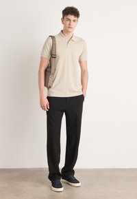 Beige polo shirt, sorte bukser og marineblå sko. Modellen bærer en brun skuldertaske. Glat stof, skræddersyet pasform, minimalistisk design.