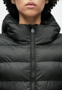 Μαύρο μπουφάν puffer με ψηλό γιακά και μεγάλο κουκούλα. Διαθέτει φερμουάρ μπροστά με ασημί λογότυπο 'HUGO'. Η υφή είναι λεία και ελαφρώς γυαλιστερή.