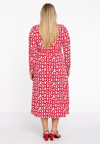 Rot-weißes, knielanges Kleid mit langen Ärmeln, das ein Blattmuster zeigt. Das Design hat eine taillierte Taille und einen ausgestellten Rock, kombiniert mit roten Absatzsandalen.