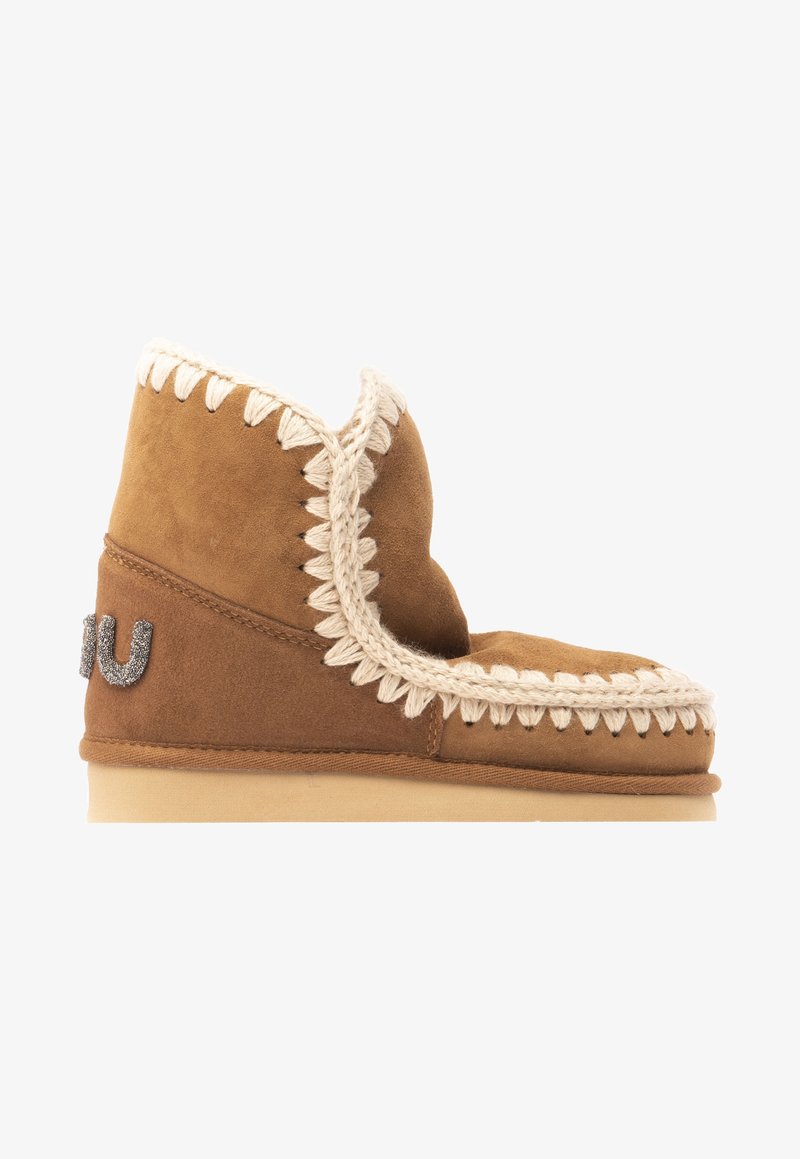 Brown suede ankle boot with a decorative cream stitching along the top edge and a metallic logo detail on the outer side. Flat sole.