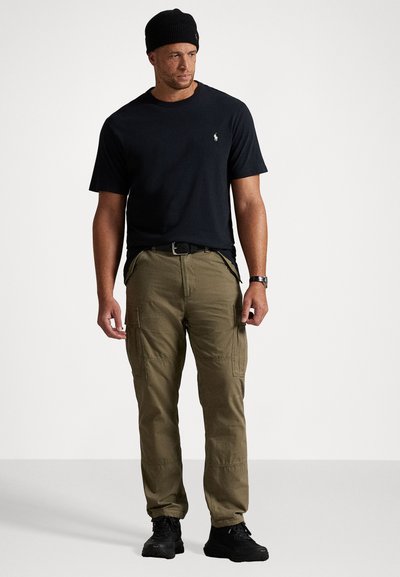 Melnas īsām piedurknēm T-krekls, olīvzaļi kravas bikses ar kabatām, valkātas ar melnu beanie cepuri un melnām kedām.