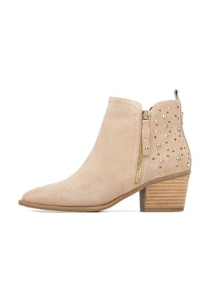Botín beige de ante con punta afilada y tacón de madera apilada. Presenta cremallera dorada y tachuelas decorativas en un lado.