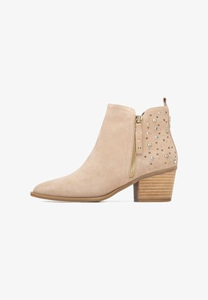 Botín beige de ante con punta afilada y tacón de madera apilada. Presenta cremallera dorada y tachuelas decorativas en un lado.