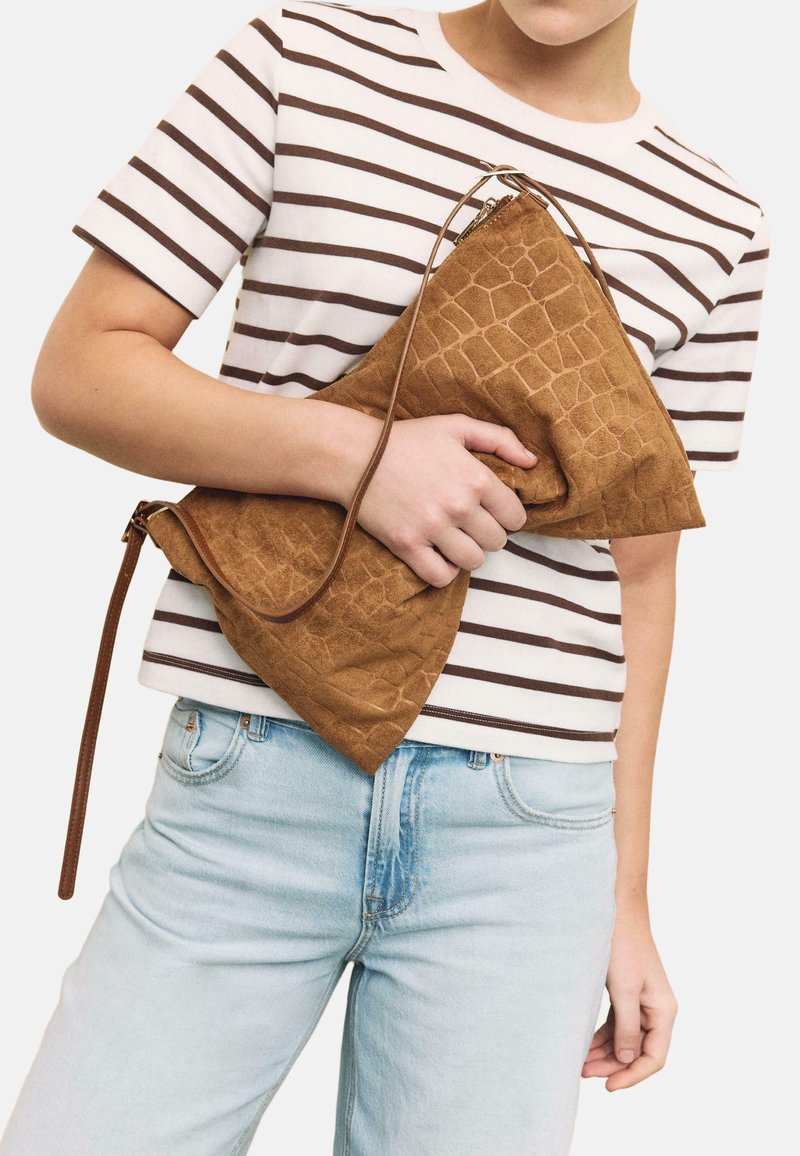 Personne portant une chemise à rayures blanches et brunes, tenant une grande pochette marron texturée avec une longue bandoulière.