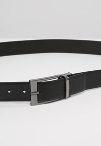 Ceinture en cuir noir avec une texture lisse, boucle métallique rectangulaire en noir mat avec un logo gravé, et plusieurs trous d'ajustement.