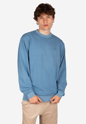 Junger Mann mit lockigem Haar trägt ein blaues Sweatshirt mit langen Ärmeln und beige Hose, steht vor einem schlichten weißen Hintergrund.