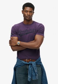 Fioletowy bawełniany t-shirt z czarną grafiką, okrągły dekolt, zestawiony z dżinsowymi spodniami i związana dżinsowa koszula w talii. Złoty detal zegarka na nadgarstku.