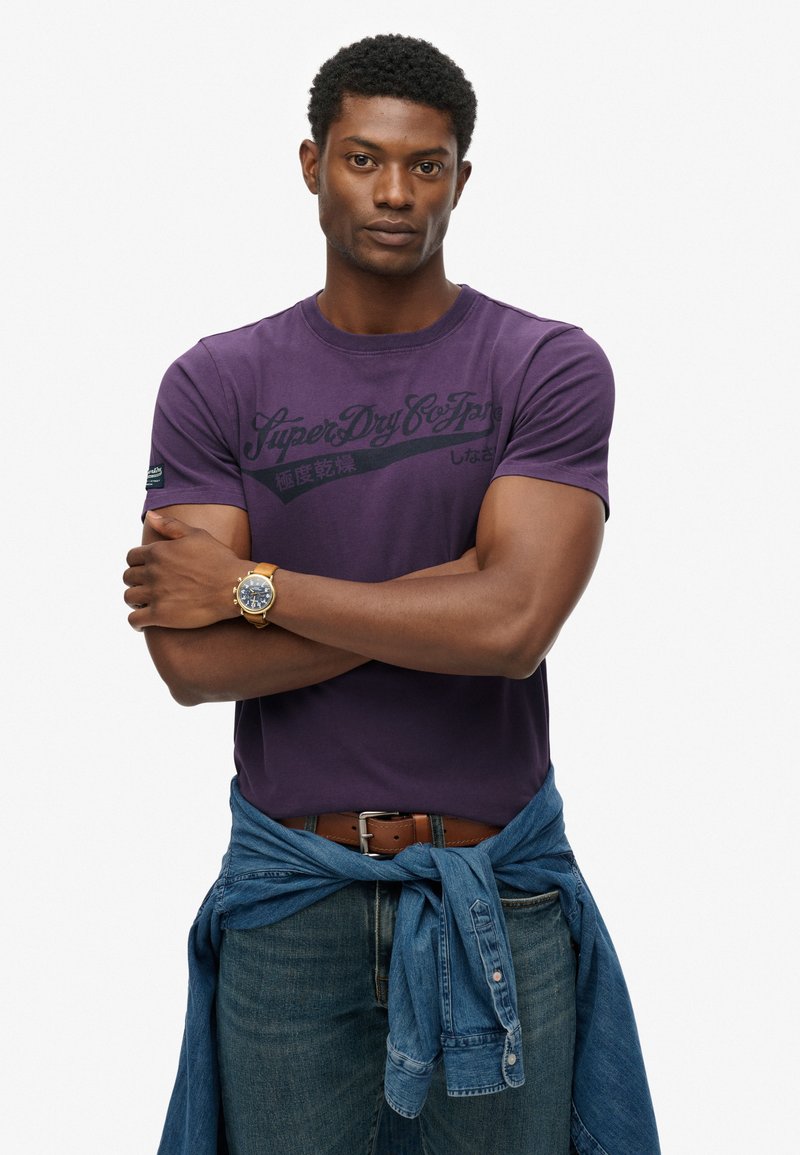 Fioletowy bawełniany t-shirt z czarną grafiką, okrągły dekolt, zestawiony z dżinsowymi spodniami i związana dżinsowa koszula w talii. Złoty detal zegarka na nadgarstku.
