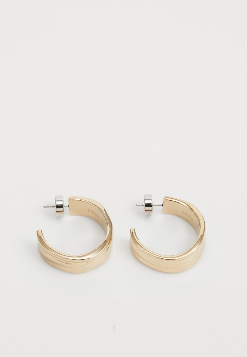 Gullige hoop-øreringer med et bølget design, som har en poleret overflate og sølvstift bak. Øreringene har en glatt, blank finish.