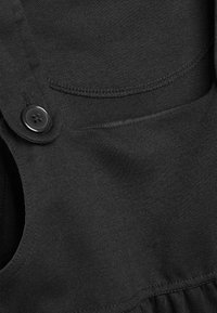 Czarna bawełniana tkanina o gładkiej strukturze, z detalem w postaci guzika oraz zaokrąglonym wzorem kieszeni. Szwów są czyste i starannie wykonane.