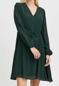 Vihreä, polvimittainen mekko, joka on valmistettu teksturoidsesta kankaasta, V-pääntiellä, pitkillä hihoilla ja kiristettävällä vyötäröllä. Ominaisuuksiin kuuluu rento, leveä siluetti.