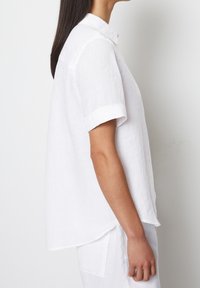 Biała lniana koszula z zapięciem na guziki, krótkimi podwiniętymi rękawami i luźnym krojem. Posiada zakrzywiony dół i teksturowany materiał.