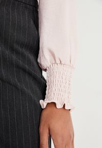 Helle rosa Bluse mit Puffärmeln und gerafften, rüschierten Bündchen, kombiniert mit einem dunkelgrauen gestreiften Rock. Die Bündchen haben eine smockte Textur.