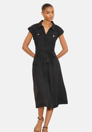Frau trägt ein schwarzes Midi-Kleid mit kurzen Ärmeln, Kragen, geknöpften Brusttaschen und Taillengürtel, steht mit Händen hinter dem Rücken.