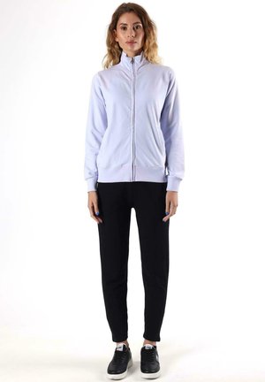 Une femme porte une veste zippée couleur lavande pâle, un pantalon noir et des baskets noires sur un fond blanc uni.