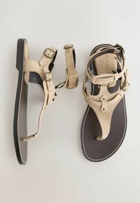 Sandales tongs en daim beige avec brides réglables à la cheville, boucles en bronze et semelles plates marron foncé avec coutures blanches.
