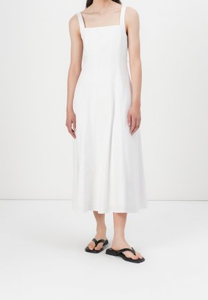 Biała lniana sukienka midi z kwadratowym dekoltem i cienkimi ramiączkami, zestawiona z czarnymi płaskimi japonkami. Prosta, czysta sylwetka.