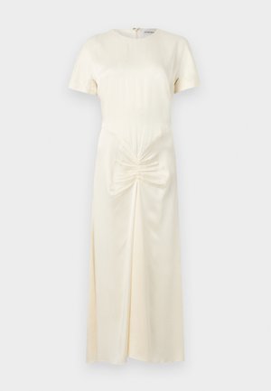 Moteris stovinti dėvi kreminės spalvos midi suknelę trumpomis rankovėmis ir raukšlėtu liemens detaliu, derinama su juodais atviri bato kulniukais su kulkšnies raišteliais.