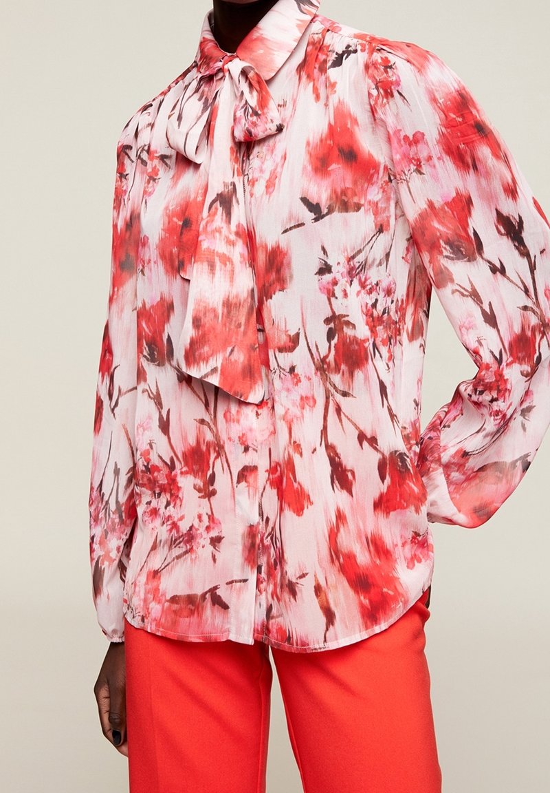Bluse mit floralem Druck aus transparentem Material, mit hohem Kragen und langen Bändern, zarten Falten und langen Ärmeln. Die Farben sind Pink, Rot und Weiß.