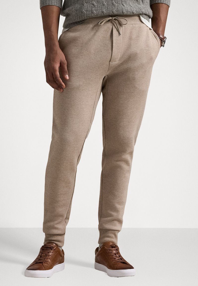 Beige jogger pants made of soft fabric, featuring an adjustable drawstring waist and side pockets, paired with brown sneakers.