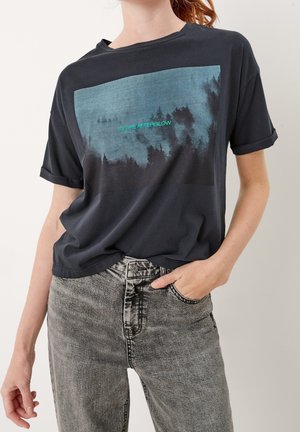 Vrouw draagt een donkergrijs T-shirt met een silhouet van een bos en de blauwe tekst "IN THE AFTERGLOW," gecombineerd met een lichtgrijze spijkerbroek.