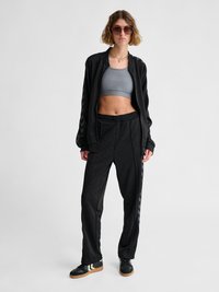 Giovane donna con tuta nera, reggiseno sportivo grigio, scarpe da ginnastica nere e occhiali da sole in piedi davanti a uno sfondo bianco uniforme.