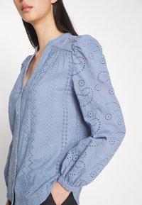 Femme portant un chemisier bleu clair à manches longues avec des motifs complexes en dentelle ajourée et de petits boutons sur le devant.