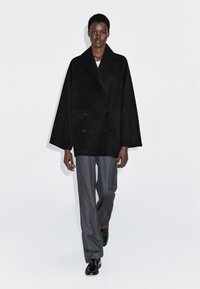 Zwarte dubbelbreasted jas met wijde mouwen en een schouderkraag, gecombineerd met grijs op maat gemaakte broeken en zwarte schoenen.