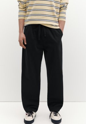 Mand iført sorte løse bukser og hvide sneakers, hænder i lommerne, beige langærmet skjorte med marineblå vandrette striber.