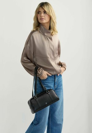 Kobieta o blond włosach nosi błyszczącą kurtkę w kolorze taupe i niebieskie dżinsy, trzymając czarną skórzaną torebkę za paski na ramię.