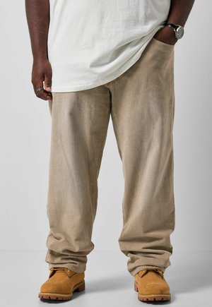 Hombre con pantalones de pana beige, botas de trabajo marrones, camisa blanca, reloj de pulsera y un anillo en la mano izquierda, de pie con una mano en el bolsillo.