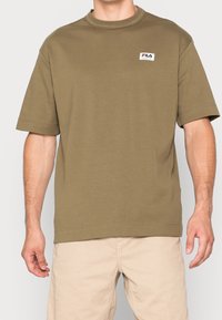 T-shirt en coton vert olive avec un col rond, des manches courtes et un petit patch logo Fila sur la poitrine. Texture lisse, coupe décontractée.