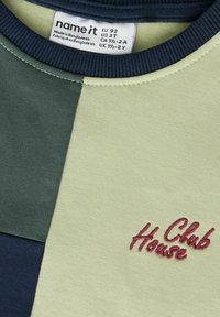 Genser i olivengrønn, marineblå og lysegrønne paneler; har ribbet hals og 'Club House' brodert i burgunder. Myk tekstur.