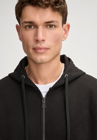 Felpa nera con zip e un motivo testurizzato, con cappuccio regolabile e un interno grigio chiaro. Finiture in metallo argentato sulla zip.