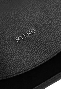 Superficie de cuero negro texturizado con el logo de la marca "RYŁKO" en metal plateado centrado en ella.