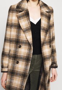 Rutig kappa i beige, brunt och krämfärgat med dubbelknäppt design, stor krage och mjukt tyg. Lager på en svart topp.