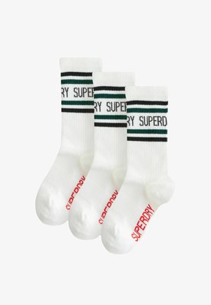 Trīs baltu zeķu pāri ar melnām un zaļām svītrām augšpusē, uz kurām ir uzdrukāts "SUPERDRY", un sarkans zīmols uz papēža.