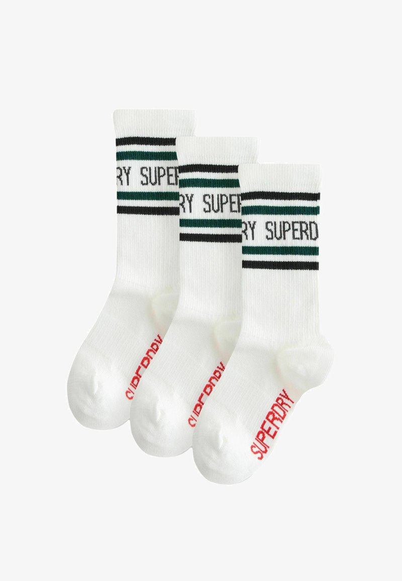 Trīs baltu zeķu pāri ar melnām un zaļām svītrām augšpusē, uz kurām ir uzdrukāts "SUPERDRY", un sarkans zīmols uz papēža.