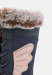 Botte d'hiver doublée en fausse fourrure avec un extérieur bleu marine, un design d'ailes pailleté rose, un détail de lacets noirs et une finition lisse et texturée.