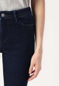 Jeans en denim foncé avec une taille haute et une légère décoloration, dotés de poches cousues et d'une fermeture à bouton en métal. Texture lisse.